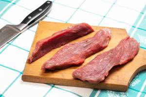 In dünne Streifen geschnittenes, rohes Rindfleisch auf Holzbrett neben Fleischmesser auf Tisch