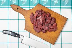 In Stücke geschnittenes rohes Rindfleisch auf Küchenbrett neben Fleischmesser auf Tisch