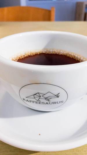 Kaffee in weißer Tasse mit Aufschrift Kaffeesaurus mit Untersetzer auf Holztisch mit Stuhl im Hintergrund
