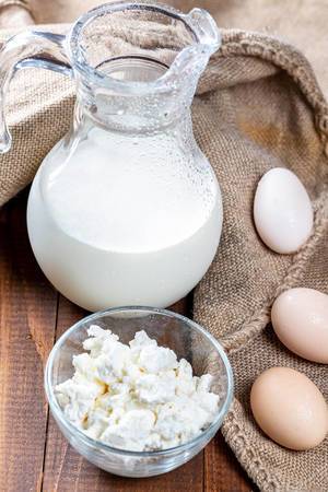 Kanne mit kalter Milch neben Glasschale mit Hüttenkäse, Eiern und Jutesack auf Holztisch