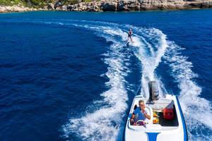 Kapitän im Wasserskiboot zieht einen Wassersportler über das tiefblaue Meer vor dem Felsenstrand von Spetses, Griechenland, als Luftaufnahme