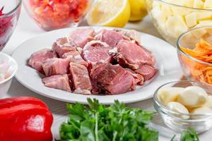 Kochrezept-Konzeptbild: Geschnittenes Fleisch, umgeben von gesunden Zutaten wie Kräutern, Knoblauchzehen, geraspelte Möhren und Kartoffeln