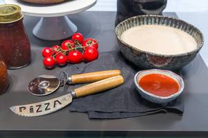 Kochutensilien für die italienische Küche: Pizzamesser, Pizzaschneider, Tomatensauce und roher Pizzateig auf einem grauen Tisch