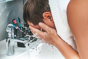 Konzept der Körperpflege: Erwachsener Mann reinigt und wäscht sein Gesicht unter laufendem Wasserhahn im Badezimmer