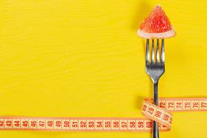 Konzept von Gewichtsverlust: Ein Stück Grapefruit auf einer Gabel, eingewickelt durch einem Bandmaß im gelben Hintergrund