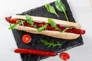 Langer Hotdog mit Räucherwurst und Salat, auf einem schwarzen Schieferstein
