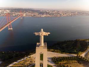 Luftaufnahme von hinten der Cristo Rei Statue über dem Tajo auf Lissabon blickend, mit Ponte 25 de Abril Brücke