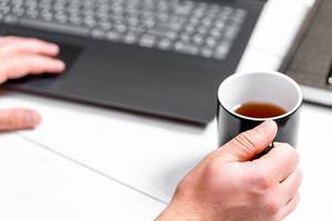 Mann erledigt Büroarbeit an Laptop mit Teetasse in der Hand