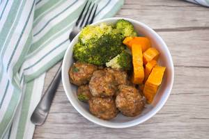Meatballs with sauce, broccoli and pumpkin in a bowl on a table with fork and kitchen towel aside