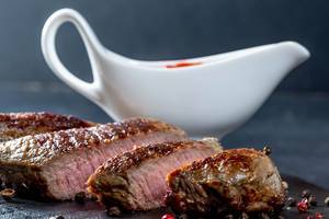 Medium gebratenes Steak in Streifen geschnitten vor weißer Sauciere vor dunklem Hintergrund