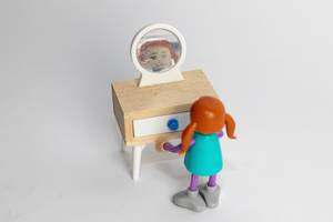 Miniatur Puppenhaus: Kleines rothaariges Mädchen mit Zöpfen sieht sich selbst in einem winzigen Tischspiegel - Nahaufnahme diagonal