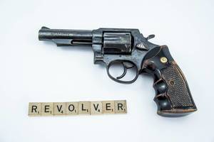 Mit Scrabble-Steinen gebildetes Wort REVOLVER vor Handfeuerwaffe auf weißem Hintergrund