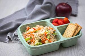 Mittagessen in einer Tupperdose - Reissalat mit Crackern und Tomaten mit einem Apfel im Hintergrund