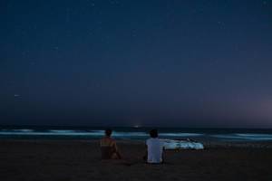 Nachtaufnahme von Frau und Mann am Strand mit Sternenhimmel, Meer und kleinem Holzboot