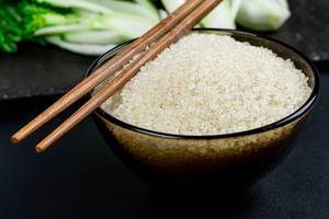 Nahaufnahme von einer Glasschüssel mit rohem Reis und Essstäbchen vor schwarzem Hintergrund