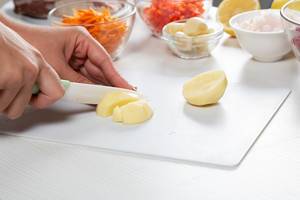 Nahaufnahme zeigt Hände beim Schneiden einer Kartoffel, mit geraspeltem und geschnittenem Gemüse im Hintergrund