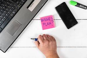 Notizzettel mit dem Text "Business Plan?" - Geschäftsplan - neben einem Handy und Laptop auf einem weißen Tisch