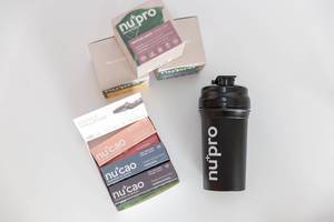 NuPro protein shakes for muscle building and raw cocoa NuCao, in various flavours, stacked on a white surface next to a drinking bottle