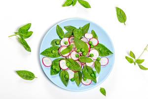 Obene Aufnahme von leichtem vegetarischem Salat aus frischen Basilikumblättern, grüne Erbsen und Radieschen auf blauem Teller