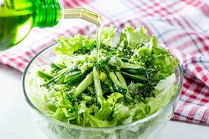 Olivenöl aus Karaffe wird über frischen grünen Salat mit Kräutern in Glasschale gegossen