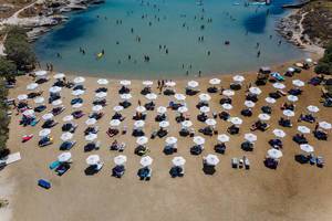 People swimming in the Mediterranean Sea and laying on beach chairs, under white parasols, at the white sandy Monastiri Beach on Paros