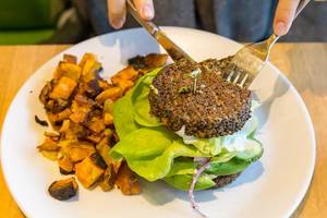 Person eating a vegan, gluten-free Inside Out Quinoa Burger (with hummus, tzatziki, organic tomato, butter lettuce, cucumber, red onion, avocado, feta) with fork and knife at the True Food Kitchen restaurant in Chicago