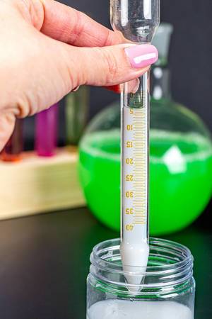 Person hält ein mit weißer Flüssigkeit gefülltes Testrohr mit Skala über ein Glas mit verschiedenen Laborgläsern im Hintergrund