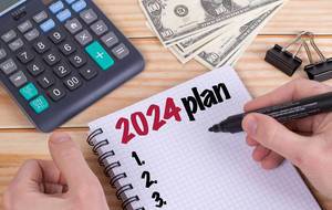 Person schreibt einen Plan für 2024 mit Filzstift in ein Notizbuch auf einem Holztisch mit Taschenrechner, Dollar Geldscheinen und Heftklammern