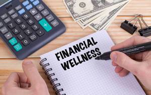 Person schreibt Text Financial Wellness mit Filzstift in ein Notizbuch auf einem Holztisch mit Taschenrechner, Dollar Geldscheinen und Heftklammern