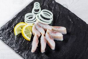 Pieces of salted herring with onion rings and lemon slices on a black stone tray