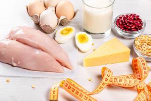 Proteinreiches Essen: Fleisch, Eier, Käse, Milch, Bohnen und Maßband auf einem weißen Tisch