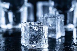Quadratische Eiswürfel mit Wassertropfen vor Wodka-Gläsern auf dunklem Hintergrund