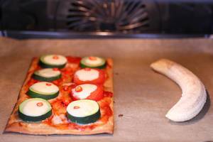 rechteckiges Pizzastück mit Zucchini- und Tomatenscheiben, überbacken mit Käse im Backofen neben einer geschälten Banane