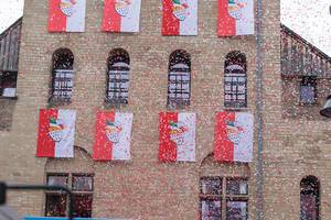 Red-white banners with the coat of arms of the city of Cologne hanging on the exterior of Severinstorburg, while the air is filled by red and white confetti during the Rose Monday parade