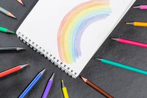 Regenbogen in weißem Notizbuch umgeben von einzelnen Buntstiften auf schwarzem Hintergrund