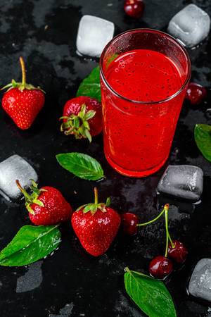 Roter Cocktail zwischen frischen Erdbeeren, Kirschen, Minzblättern und Eiswürfel, auf einem schwarzen Untergrund