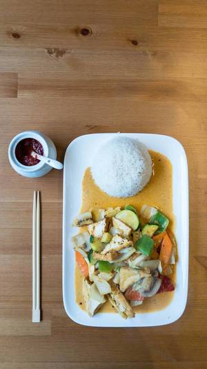 Rotes Thai-Curry mit veganen Tofu-Ecken, Zucchini, Pilzen, Paprika und Karotten auf einem Teller mit Reis, Essstäbchen und einem Gefäß mit Sambal Oelek auf einem Tisch in der Aufsicht