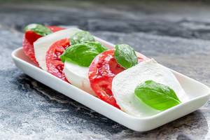 Runde Mozarellascheiben liegen mit reifer Tomate abwechselnd auf einem Teller, garniert mit grünen Kräuterblättern