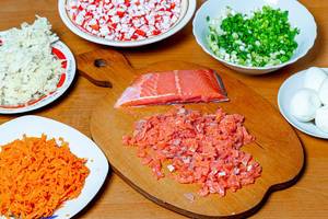 Salatzutaten wie geriebene Karotten, Kohl, Frühlingszwiebeln, gewürfelter Lachs und Krabben-Sticks auf Tellern, Schüsseln und Schneidebrett