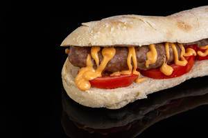 Sandwich gefüllt mit Grillwurst, Tomaten und Senf vor spiegelndem schwarzem Hintergrund