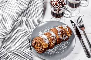Schokoladen-Biskuit-Rolle mit Kokosraspeln, neben einem Kaffee, auf einem weißen Tisch im Landhausstil