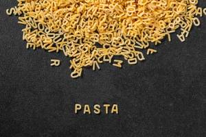 Schriftzug Pasta aus zu Buchstaben geformten Nudeln vor schwarzem Hintergrund mit weiteren Nudeln