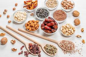 Set nuts, seeds, dried fruits in different bowls and wooden spoons on white wooden background