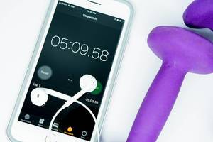 Smartphone zeigt Fitness-Stoppuhr, darauf liegen Kopfhörer, daneben violette Hanteln