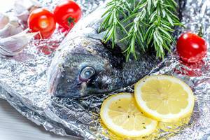 Speisefisch grillen: Nahaufnahme eines Dorade-Fischs /Goldbrasse mit Rosmarin, Zitronenscheiben und Tomaten auf Alufolie