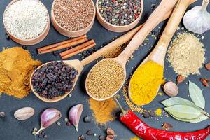 Spices - curry and mustard seeds on wooden spoons with coloured pepper seeds, flax seeds ans sesame in wooden bowls, cinnamon, garlic and nutmeg on black background