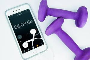 Sport-Timer auf Mobiltelefon mit Kopfhörern, daneben zwei violette Fitnessgewichte
