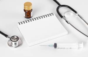 Stethoskop, Spritze, Medikamente und leeres Notizbuch auf einem weißem Tisch