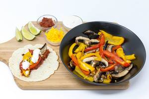 The Hello Fresh vegan fajita with peppers and muschrooms, with hot tomatosauce and lime-cream and cheddar cheese on a wooden board and the ingredients on white background