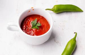 Tomato Sauce in a Cup standing next to two green Chili Peppers on white wooden table